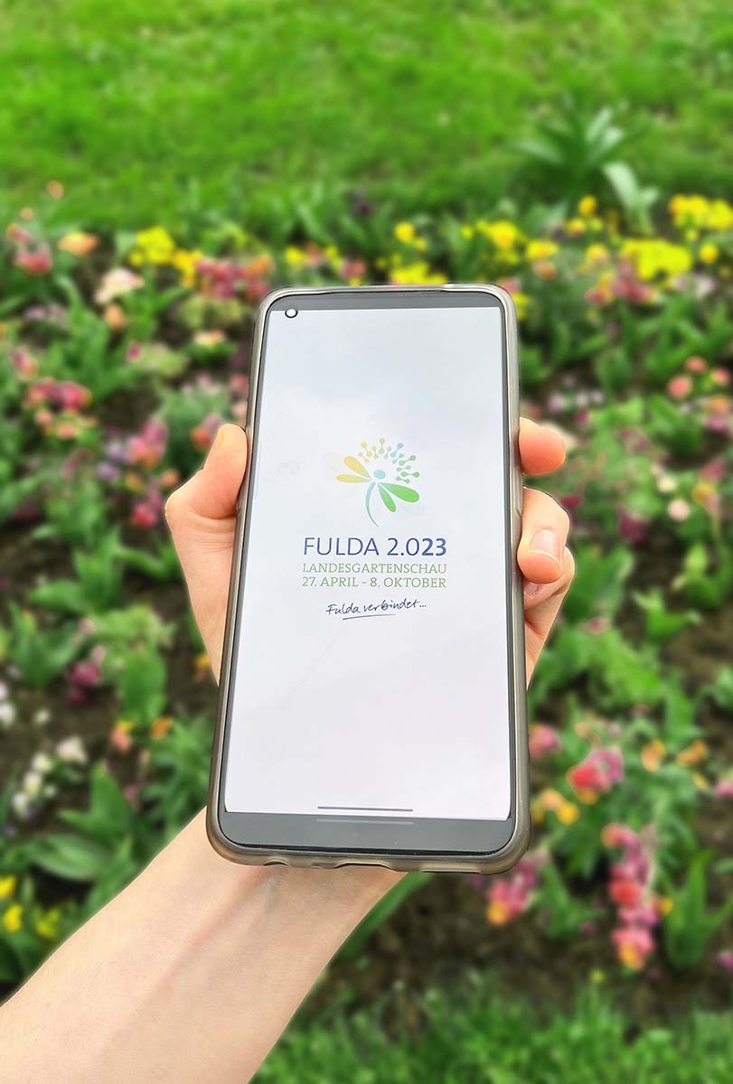 Hand, welches Smartphone hält und den Startscreen der Gartenschauapp anzeigt. im Hintergrund ist Gras und ein Blumenbeet zu sehen.
