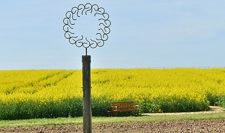 in einem Kreis zusammengeschweißte Agrarwerkzeuge auf einem Holzpfosten vor einem Rapsfeld - Kunstwerk von Claus Weber