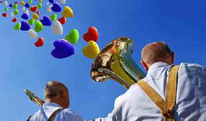 zwei Musiker mit Blechblasinstrumenten. Aus der Tuba fliegen bunte Herzen in den blauen Himmel