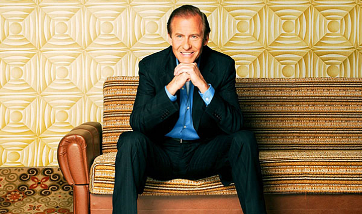 Michael Holm im Jaquett, der auf einem Sofa sitzt. Teppich Tapete und Sofa sind im 80er Jahre Stil.