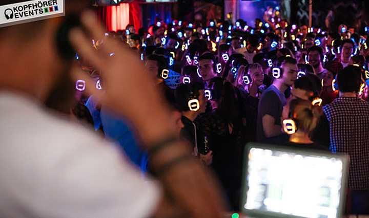 DJ mit Laptop schaut auf eine Menschenmenge tanzender Personen mit bunt leuchtenden Kopfhörern
