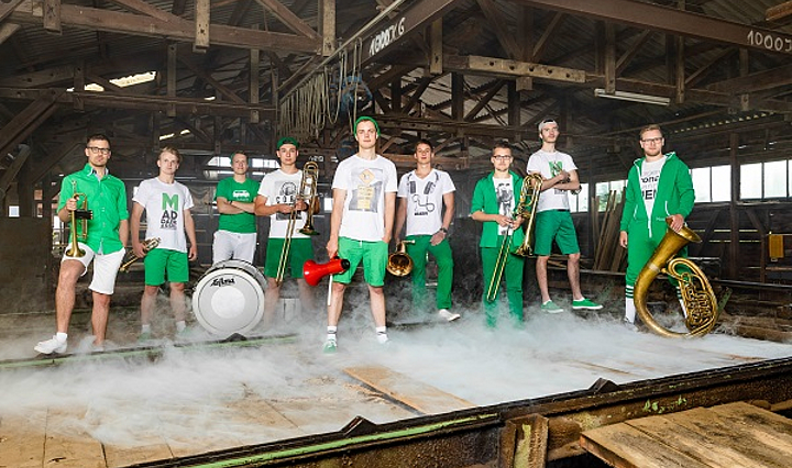 Mitglieder der Band MaddaBrassKa in grüner Kleidung mit Instrumenten in einer Scheune 